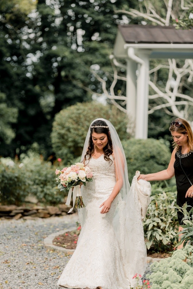 A bride walking outdoors with an assistant holding her train